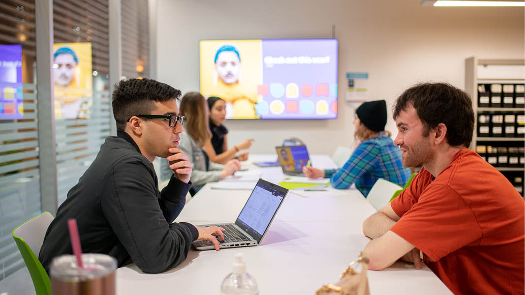 Students seated at tables with computers, discussing their learning